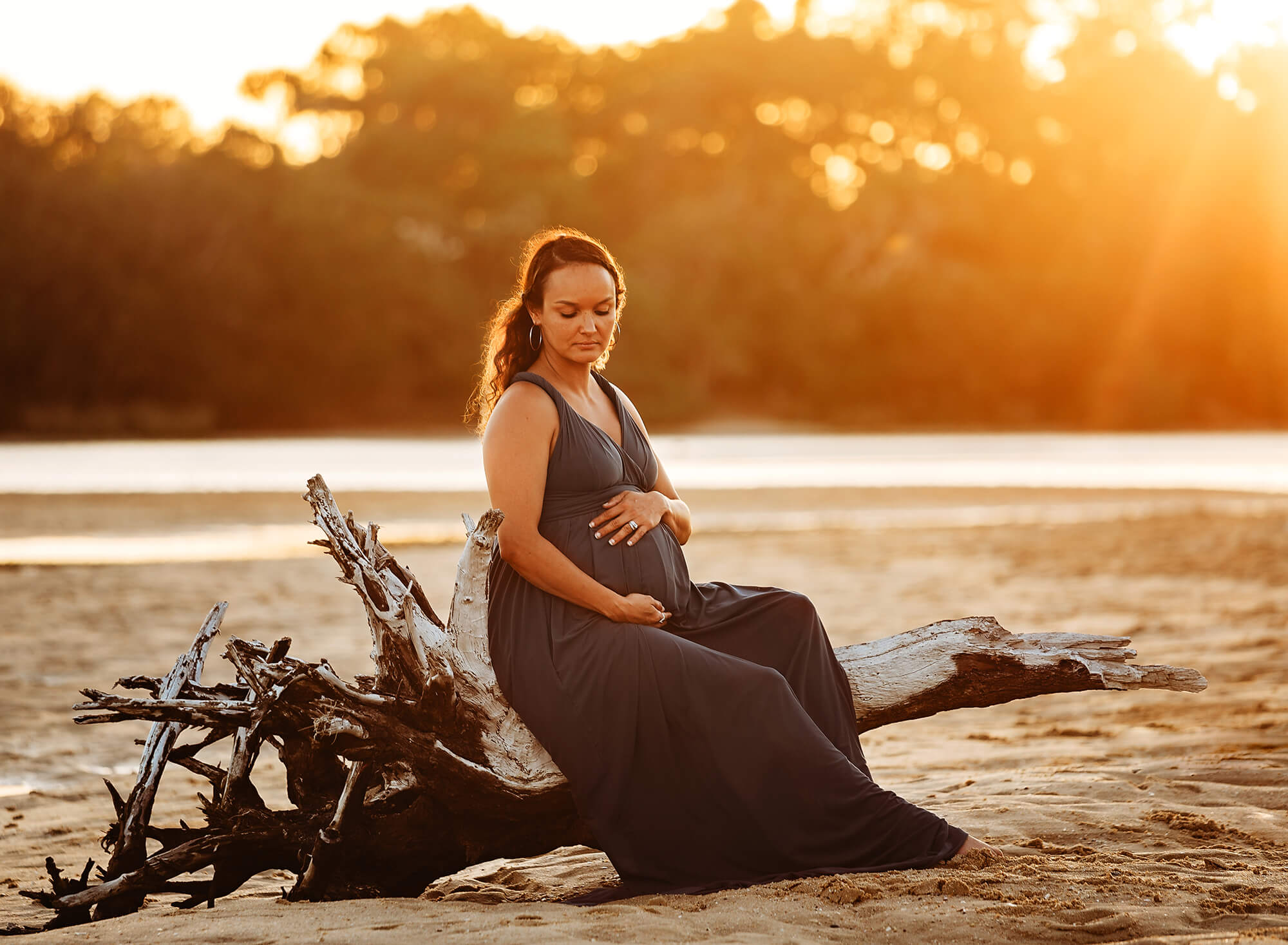 maternity photo shoot of a pregnant mother posing on a broken tree at currimundi lake queensland with the warm summer sunset behind her.