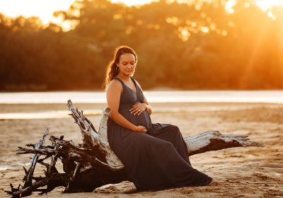 maternity photo shoot of a pregnant mother posing on a broken tree at currimundi lake queensland with the warm summer sunset behind her.